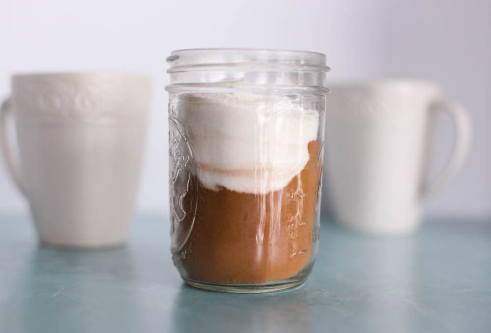 Small mason jar of homemade hot chocolate mix with two mugs in the background.