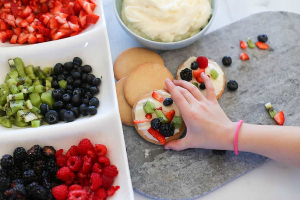 Hand grabbing a fruit pizza cookie with dishes of fruit on the table.