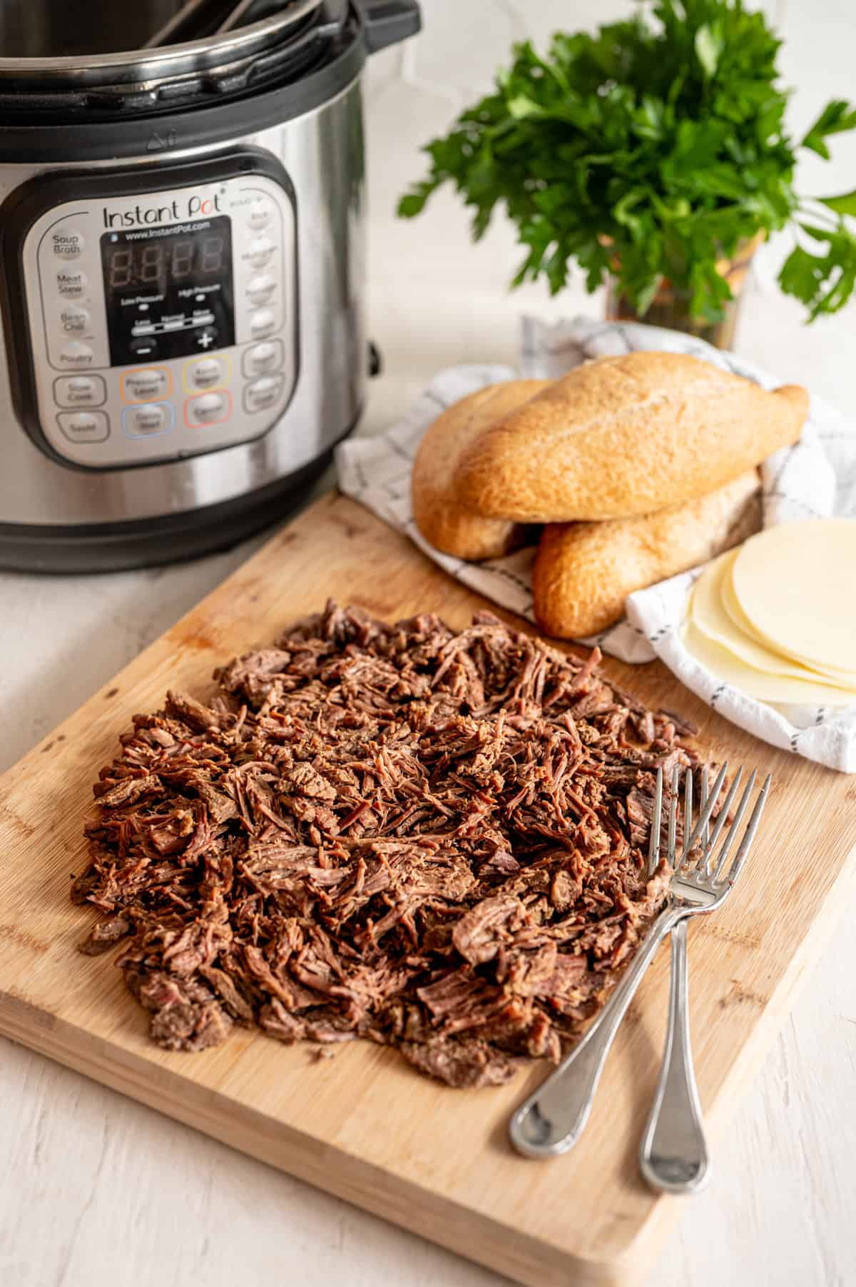 French dip shredded roast in front of an Instant Pot with ciabatta rolls and provolone slices on the side.