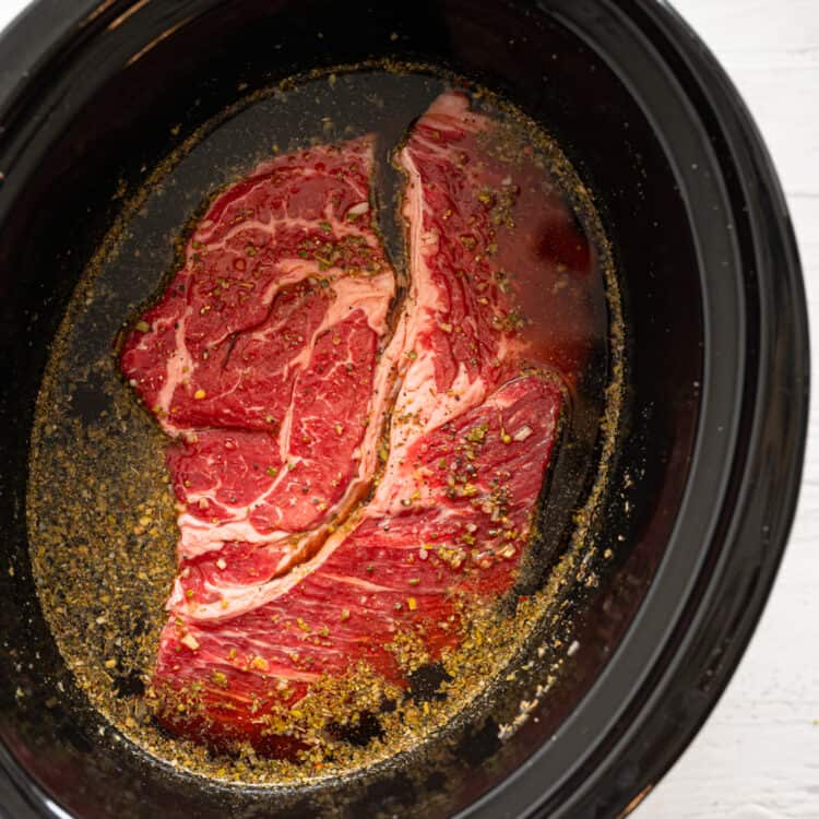 Raw chuck roast in a crock pot surrounded by French dip sandwich ingredients.