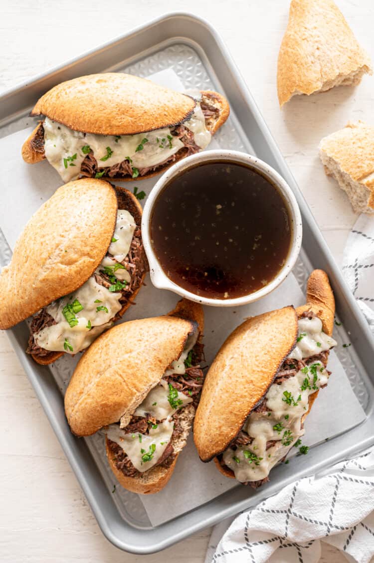 French dip sandwiches on a baking sheet surrounding a bowl of au jus.