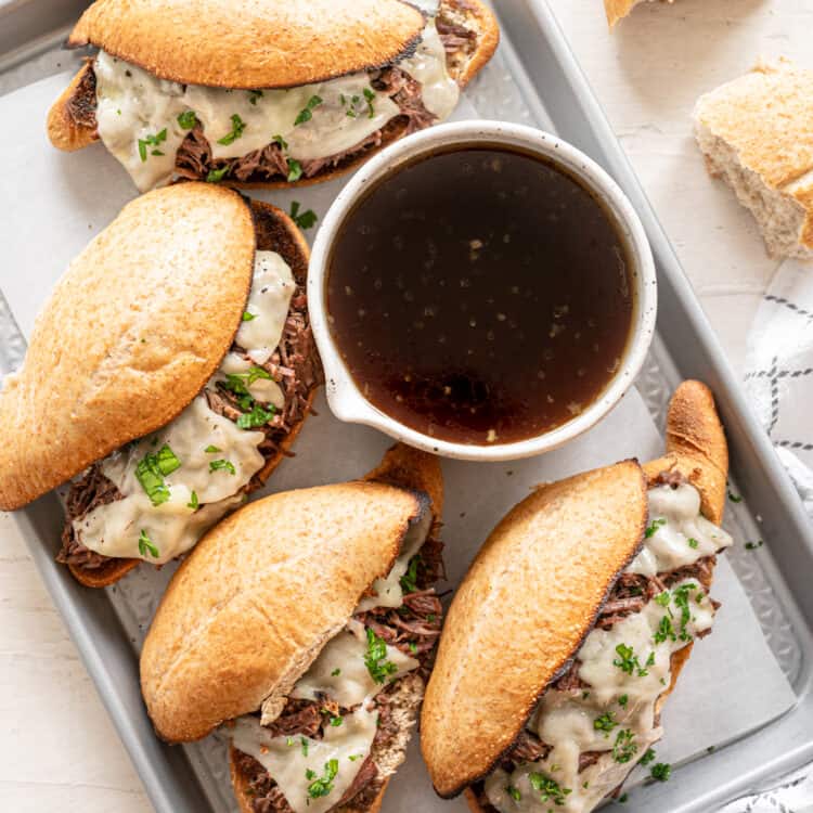 French dip sandwiches on a baking sheet surrounding a bowl of au jus.