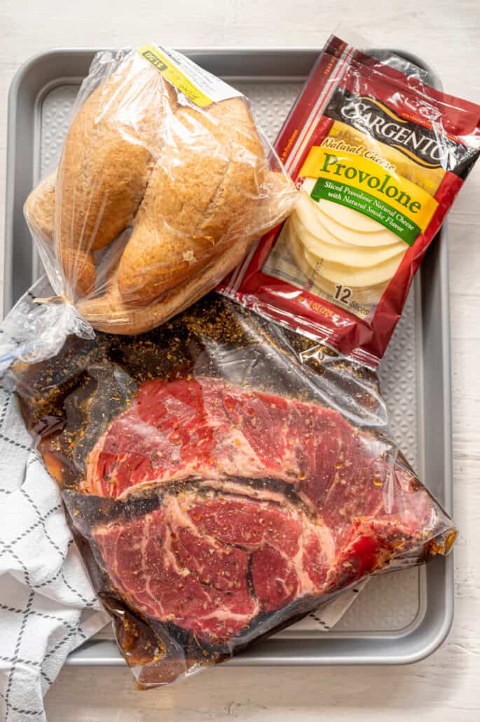 French dip freezer meal kit with marinated roast in a freezer bag, ciabatta rolls in a bag and a package of sliced provolone all sitting on a baking sheet.
