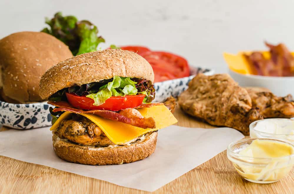 Grilled chicken sandwich with bacon, lettuce, tomato, and cheese on a cutting board.