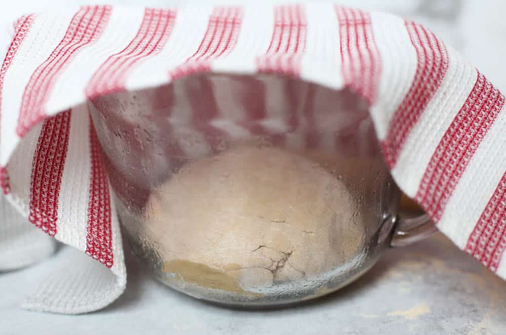 Whole wheat pizza dough rising in a glass bowl with a tea towel over top of it.