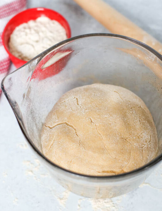 Pizza dough ball in a glass mixing bowl with a small bowl of flour and a rolling pin next to it.