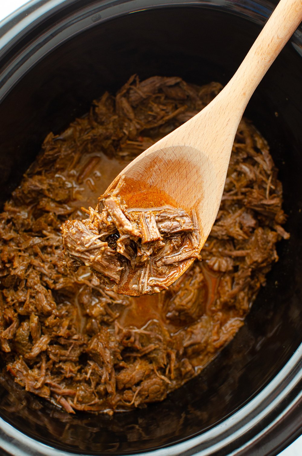 BBQ shredded beef in the slow cooker being stirred with a wooden spoon.