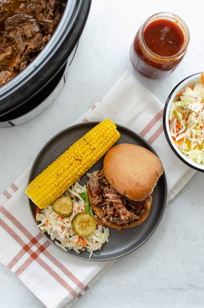 A bbq beef sandwich on a grey plate with sides of cole slaw and corn on the cob. 