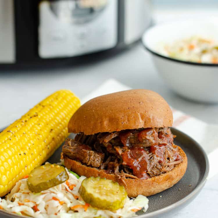 BBQ shredded beef on a bun with corn on the cob and coleslaw on a plate.