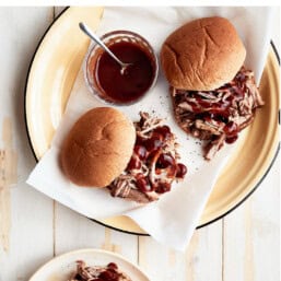 Two pulled pork sandwiches on a plate with a small bowl of BBQ sauce.