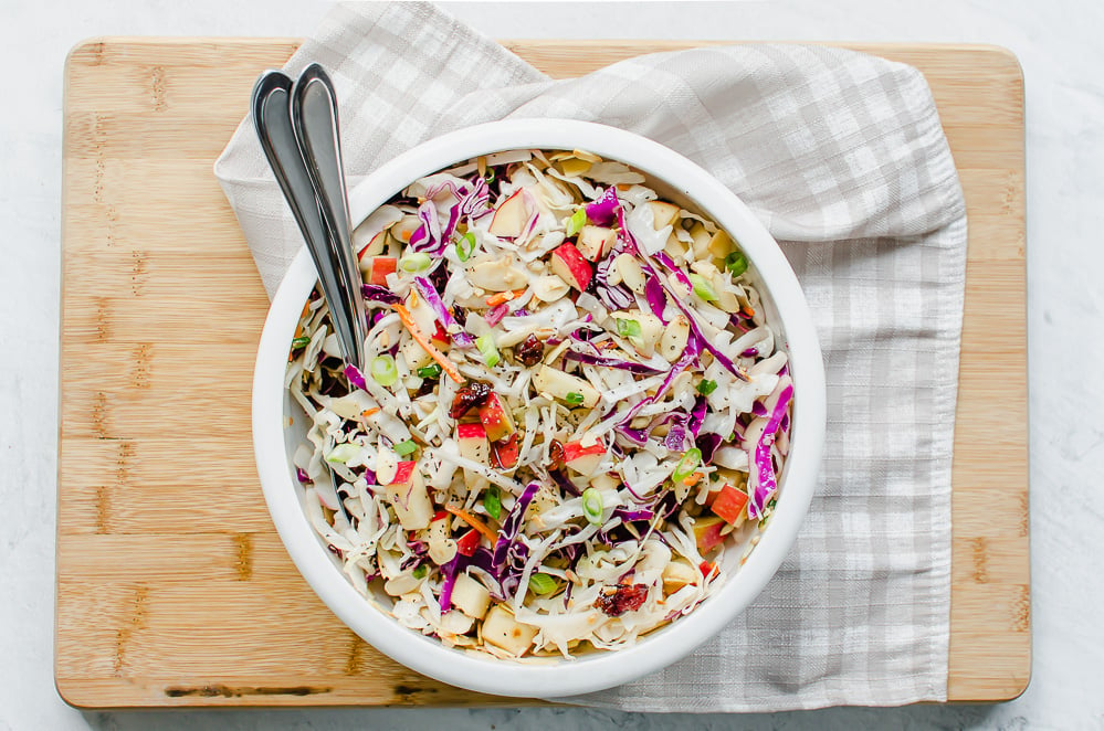 A white bowl filled with Asian slaw on a cutting board with serving utensils and a gingham towel on the side. 