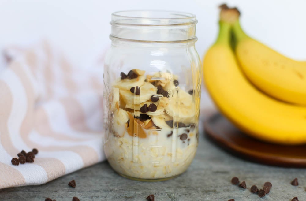 Peanut butter cup overnight oats in a mason jar on a counter with whole bananas in the background and mini chocolate chips sprinkled around the jar.