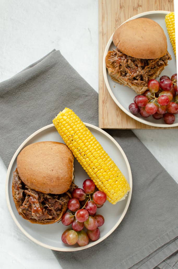 BBQ shredded beef sandwich on plates with corn and grapes