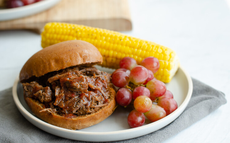BBQ shredded beef sandwich on a plate with corn and grapes