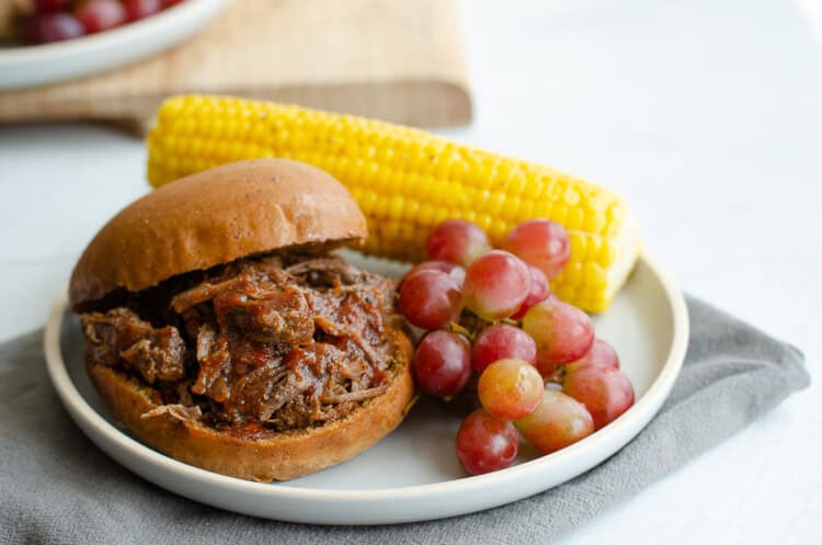 BBQ shredded beef sandwich on a plate with corn on the cob and grapes.