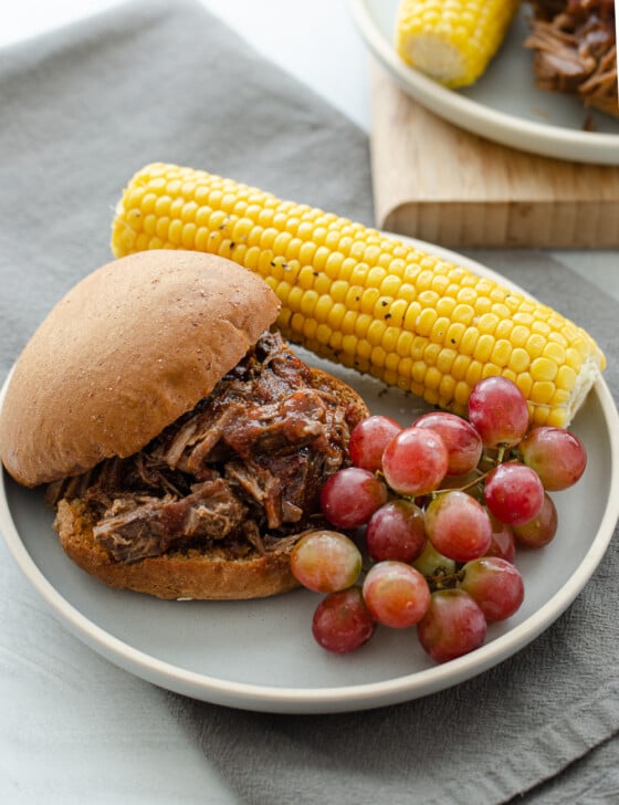 BBQ shredded beef sandwich on a plate with corn on the cob and grapes.