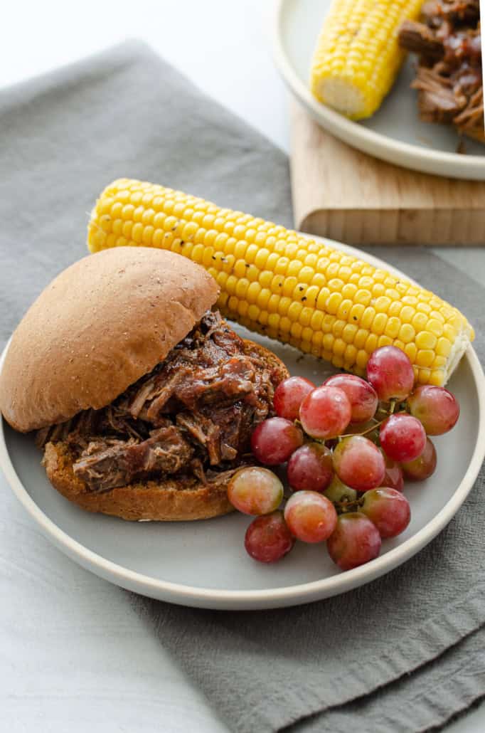 BBQ shredded beef sandwich on a plate with corn and grapes