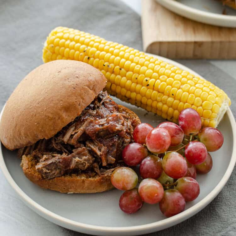 BBQ shredded beef sandwich on a plate with corn on the cob and grapes.