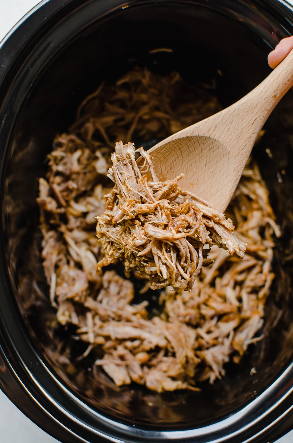Cooked pulled pork on a wooden spoon coming out of the slow cooker.