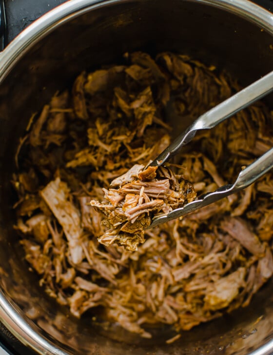 Shredded Pulled Pork in an instant pot with tongs holding a serving.