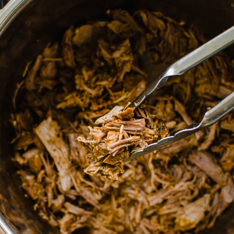Shredded Pulled Pork in an instant pot with tongs holding a serving.