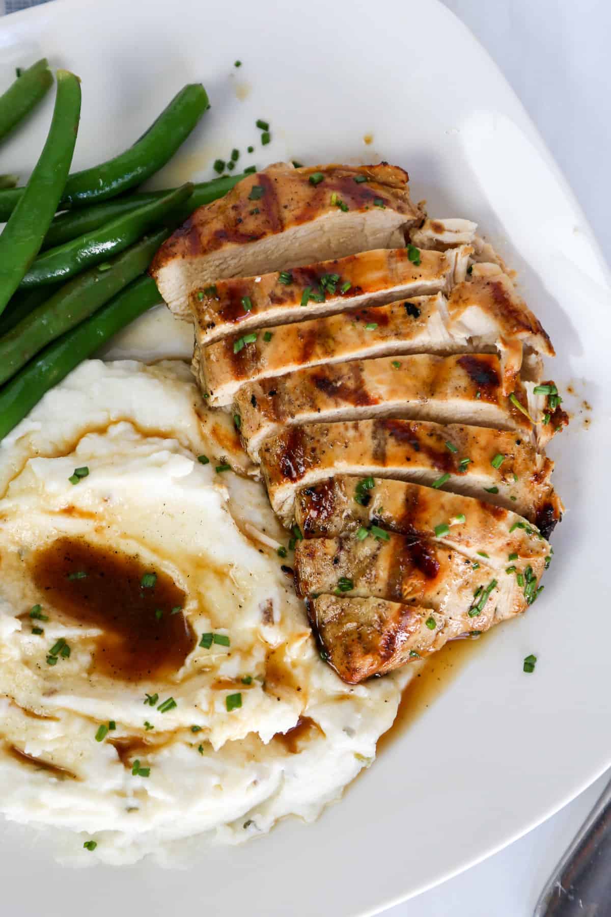 Grilled savory chicken breast sliced with mashed potatoes and green beans on a plate.