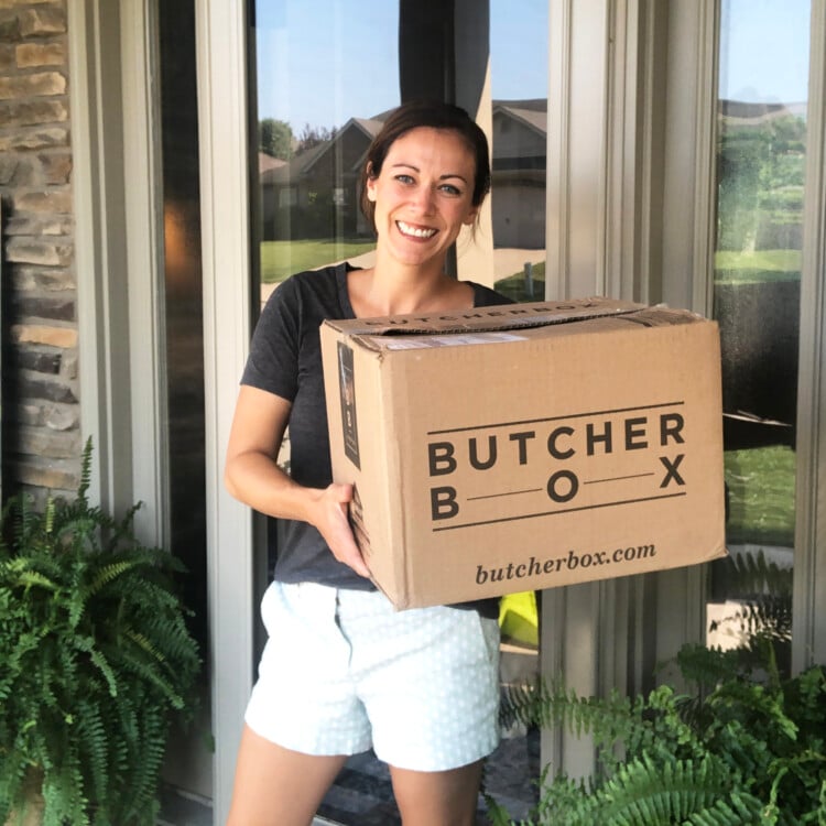 A woman standing on her front porch holding a box from Butcher Box meats.
