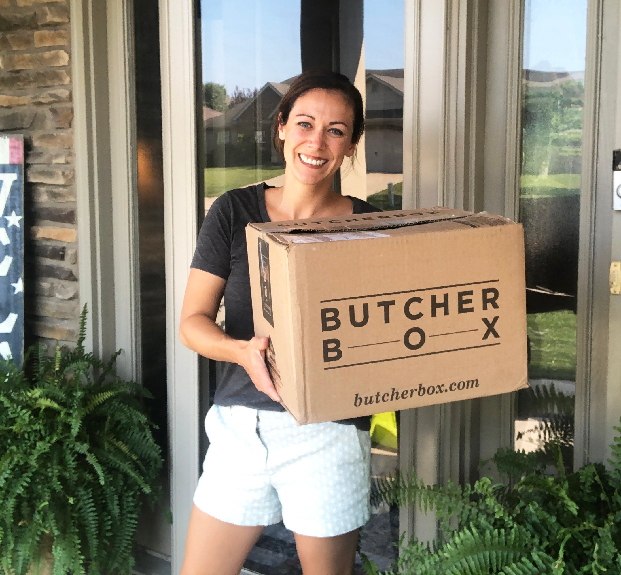 A woman standing on the porch holding a shipment from Butcher Box.