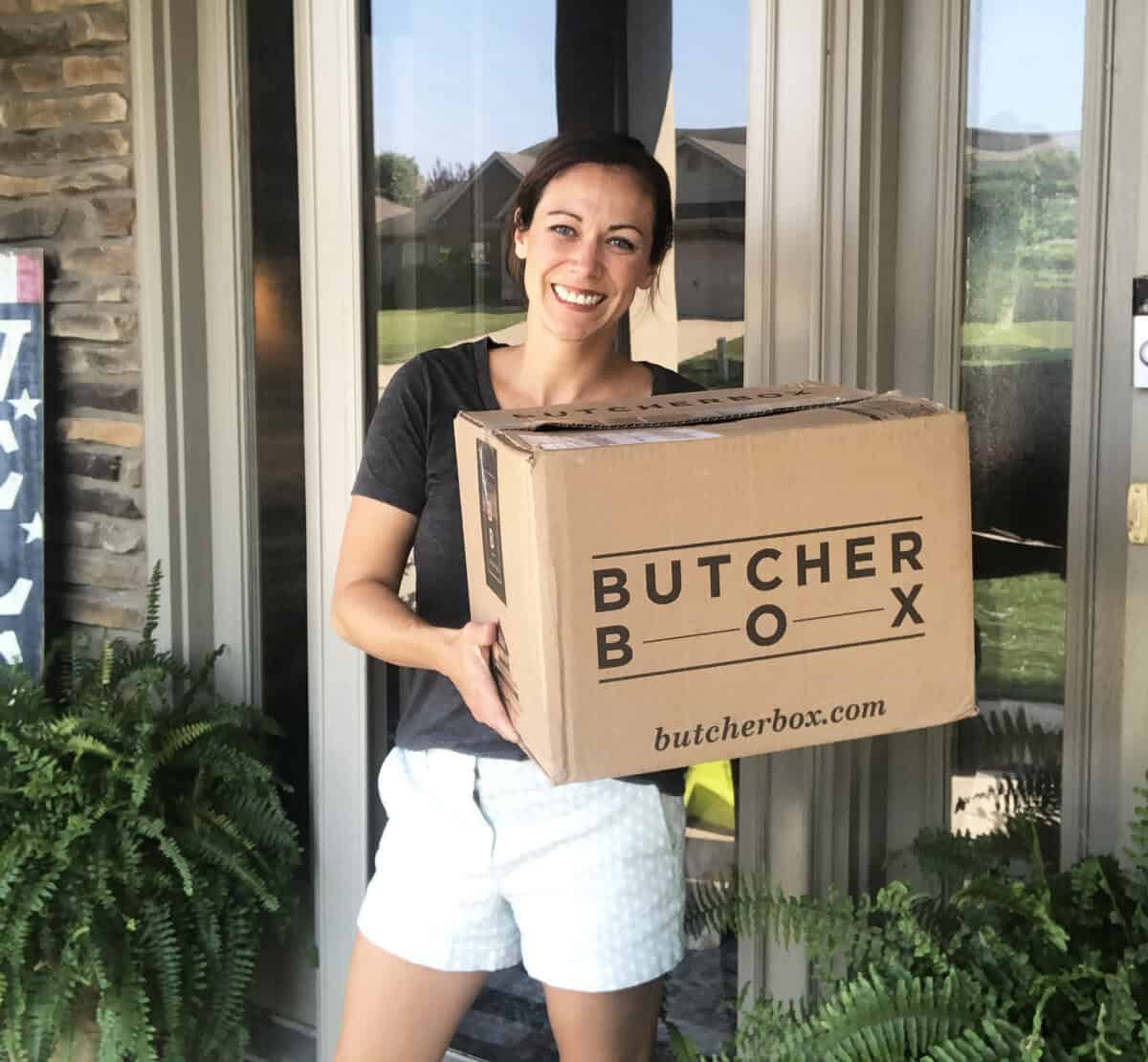 A woman standing on her front porch holding a box from Butcher Box meats.