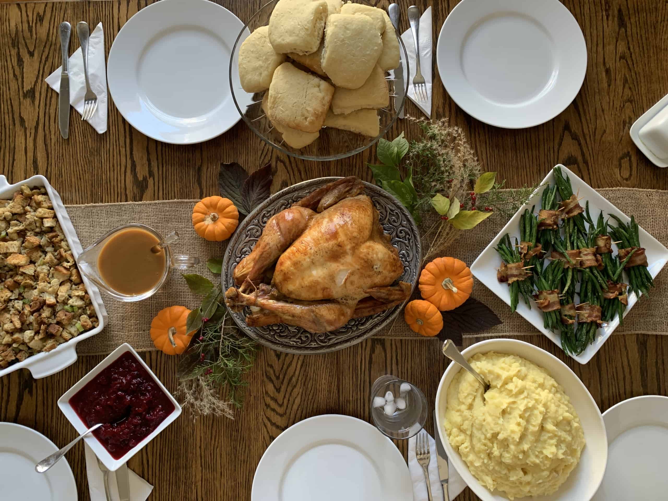 Thanksgiving table setting with turkey in the center and surrounded by mashed potatoes, green beans, stuffing, and cranberry sauce.