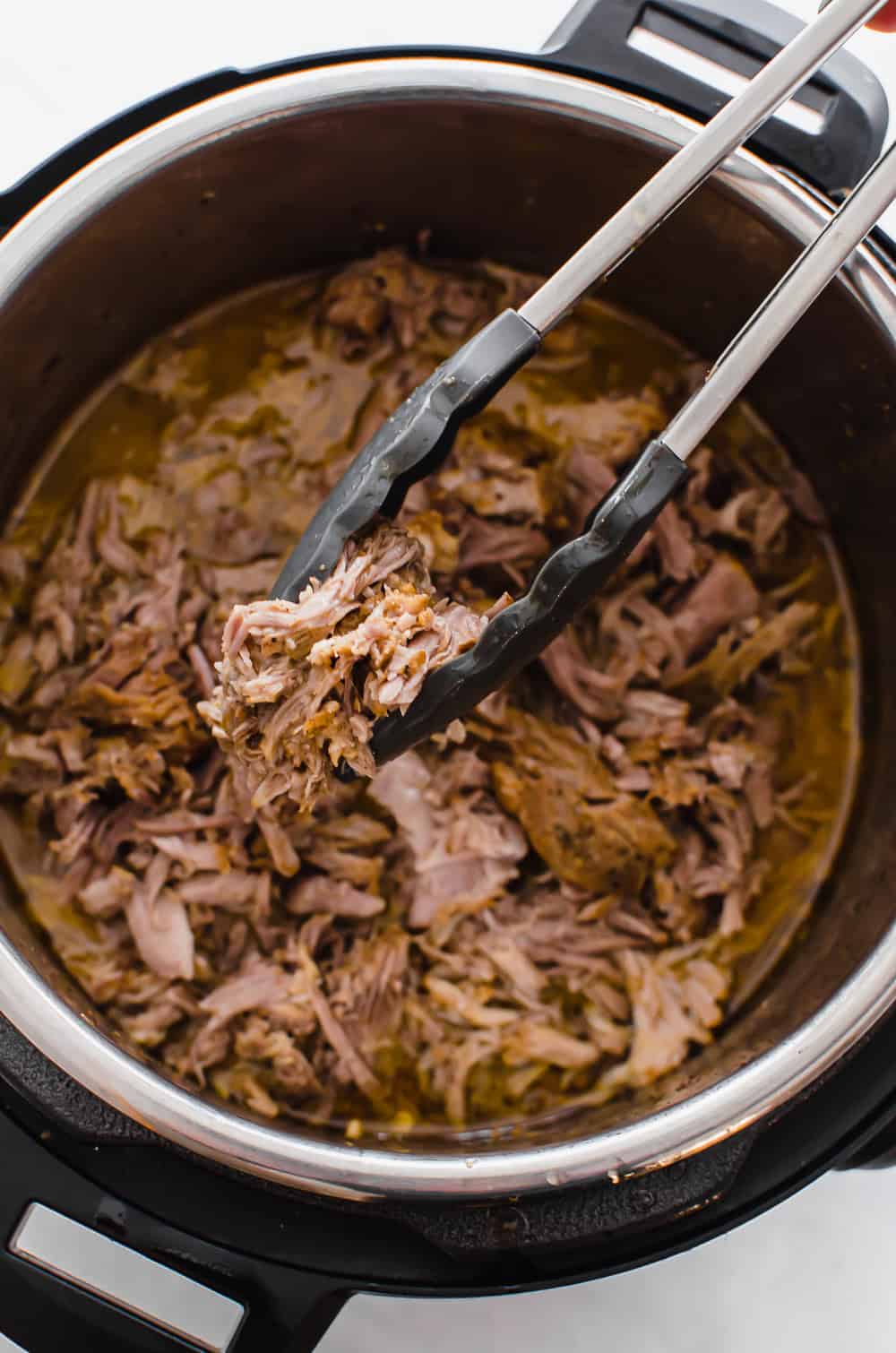 Shredded pork in an Instant Pot with tongs pulling some out.