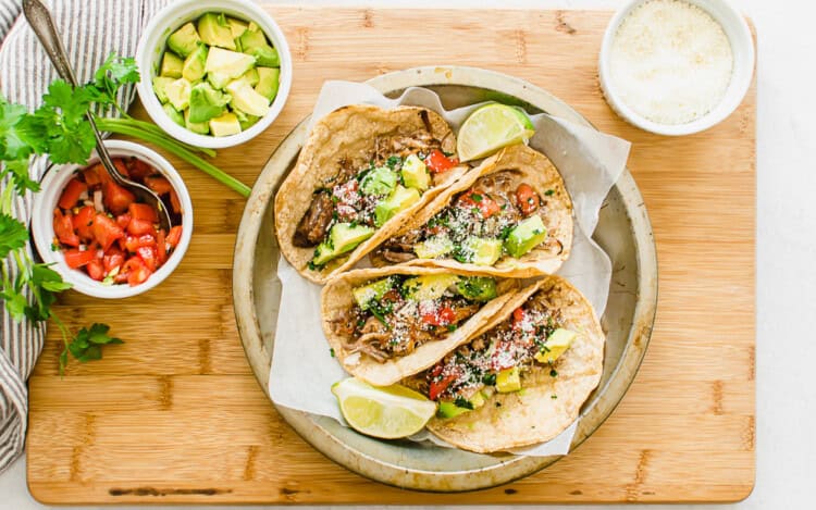 Three carnitas tacos on a plate with toppings in bowls in the background.