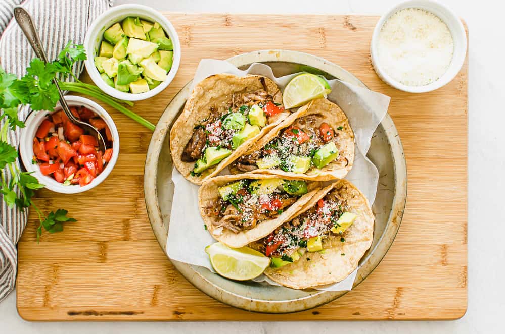 Three carnitas tacos on a plate with toppings in bowls in the background.