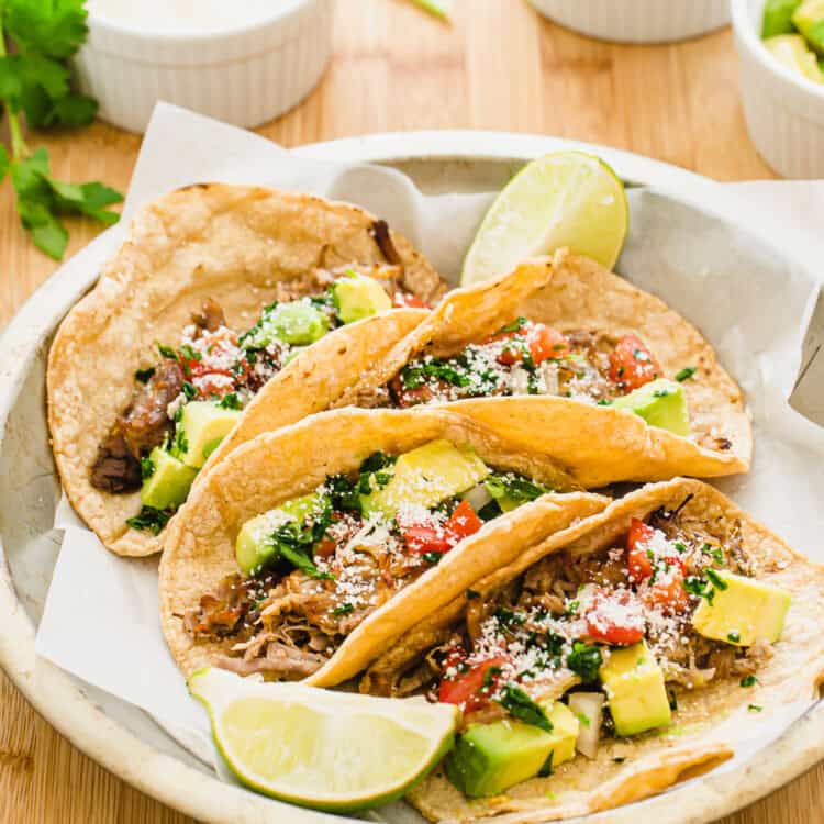 Crispy carnitas tacos in a metal dish with white bowls of toppings on the side.