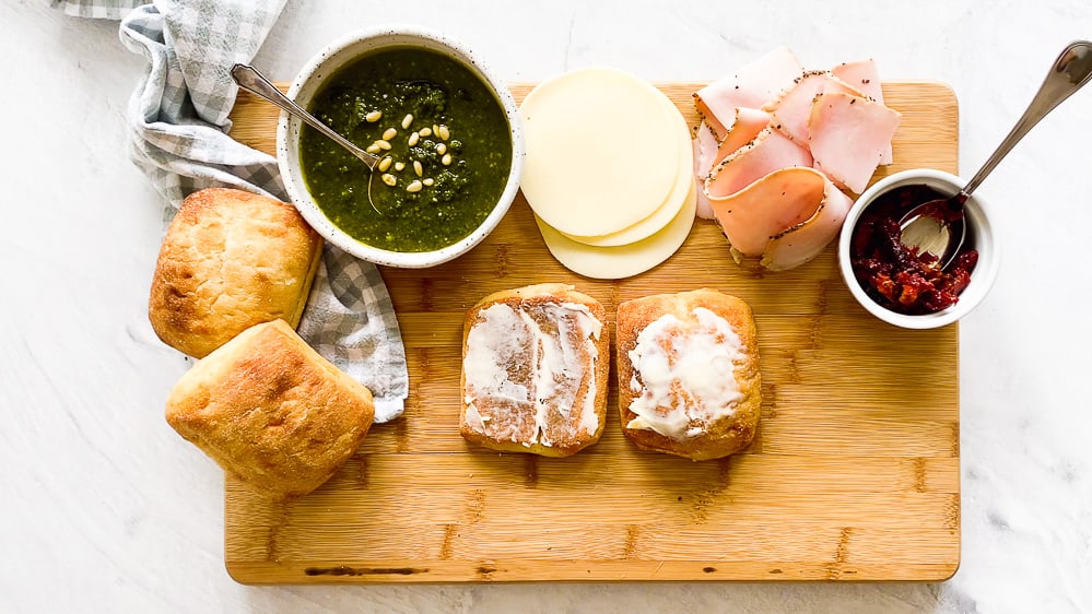 Turkey pesto panini ingredients on a wooden cutting board with the ciabatta rolls buttered in the middle.