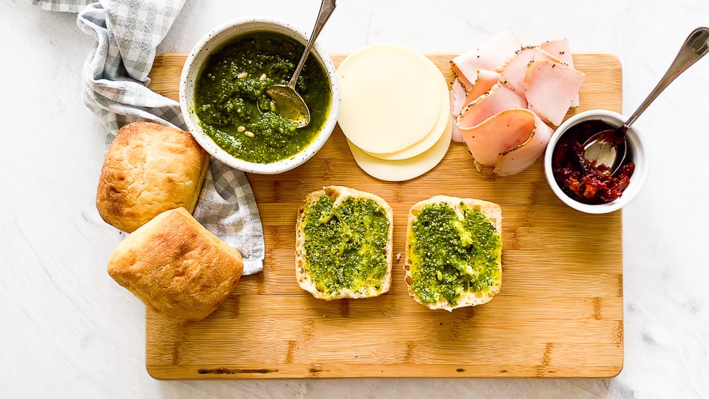 Turkey pesto panini ingredients on a wooden cutting board with the ciabatta rolls slathered with pesto in the middle.