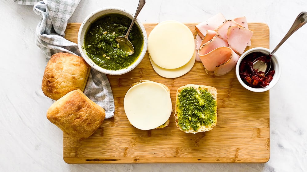 Turkey pesto panini ingredients on a wooden cutting board with ciabatta roll halves in the middle with pesto and provolone cheese on it.