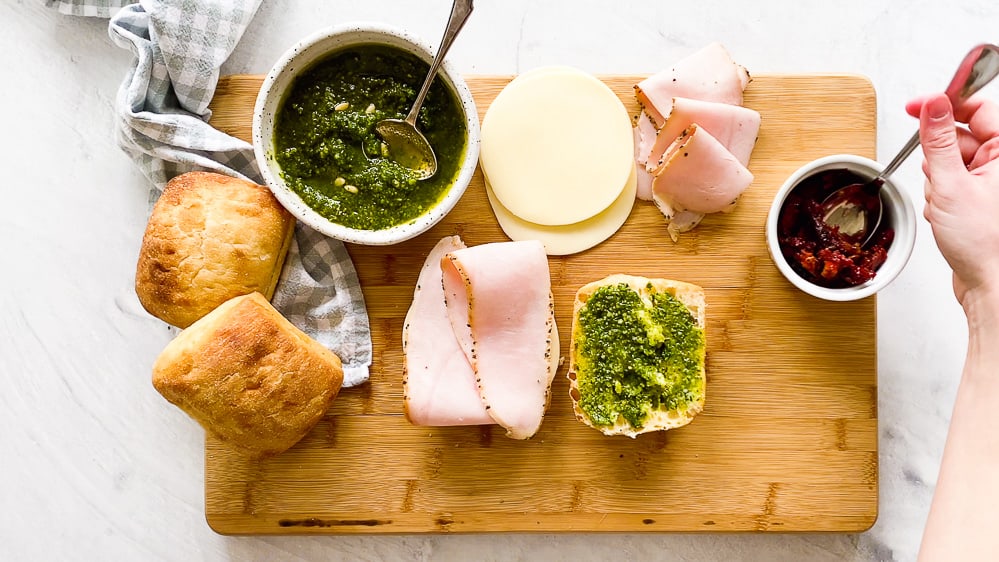 Turkey pesto panini ingredients on a wooden cutting board with ciabatta roll halves in the middle with pesto on one half and provolone cheese and turkey on the other.