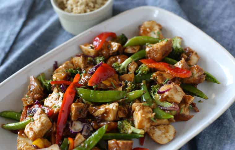 Honey bourbon chicken served with stir fry veggies on a white platter with brown rice on the side.