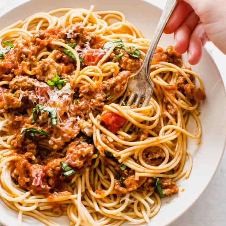 A hand with a fork twirling a bite of spaghetti on a plate.