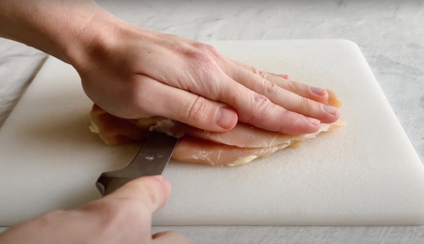 Two hands slicing a chicken breast lengthwise on a cutting board.