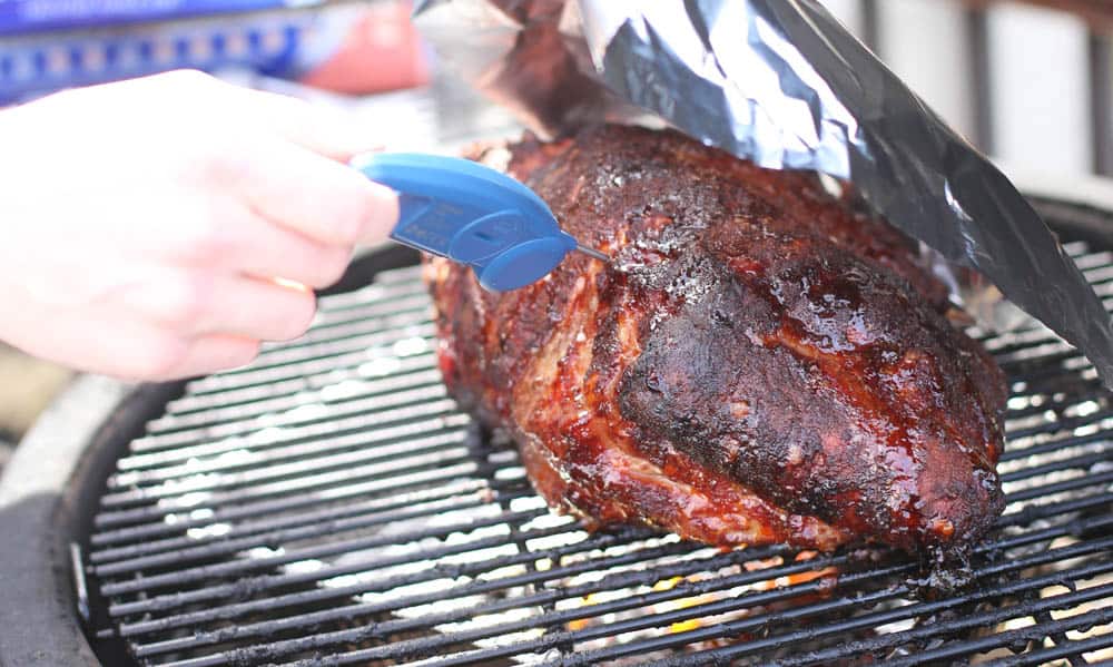 Checking the internal temperature of a smoked pork shoulder on the grate of a smoker.