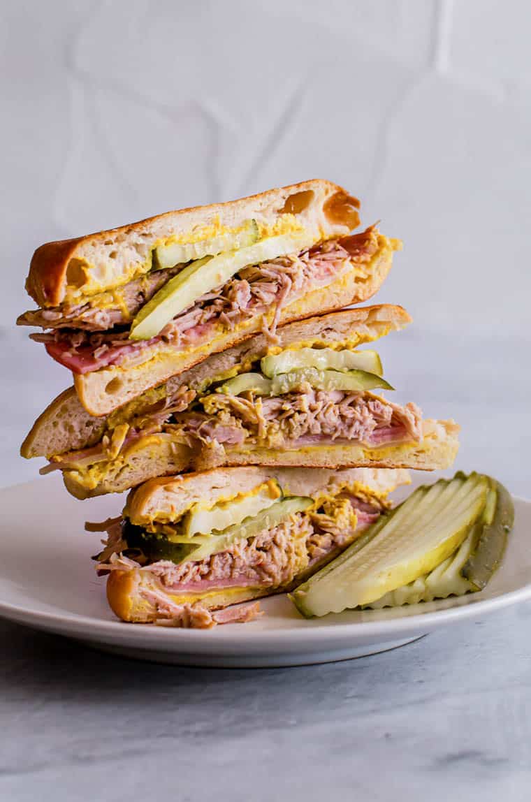 Two Cuban Panini sandwiches cut in half and stacked on a plate with extra pickle slices.