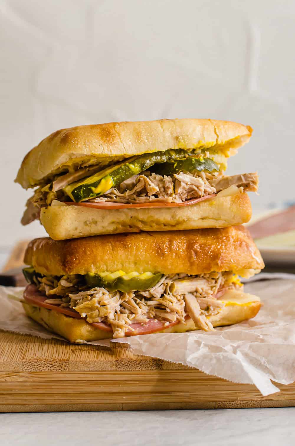 Two cuban panini sandwiches stacked on top of each other on a cutting board with a piece of wax paper. 