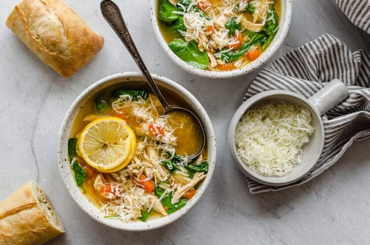 Two white bowls of lemon chicken orzo soup with crusty bread on the side.