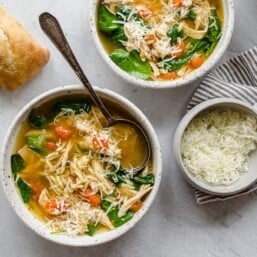 Two white bowls of lemon chicken orzo soup with spoons and crusty bread on the side.