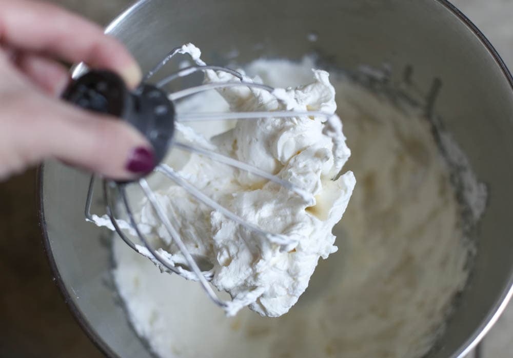 Whisk attachment of a stand mixer full of homemade whipped cream being held above the mixer bowl with more whipped cream.