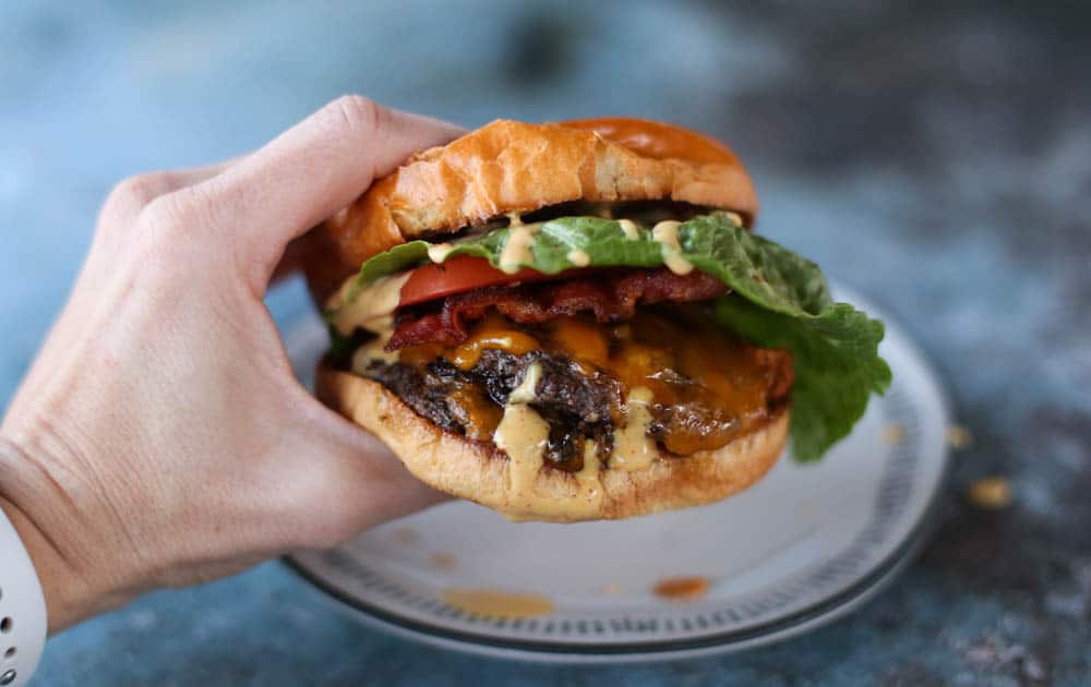 Hand holding a smash burger with lettuce, tomato, bacon, cheese, and secret sauce.