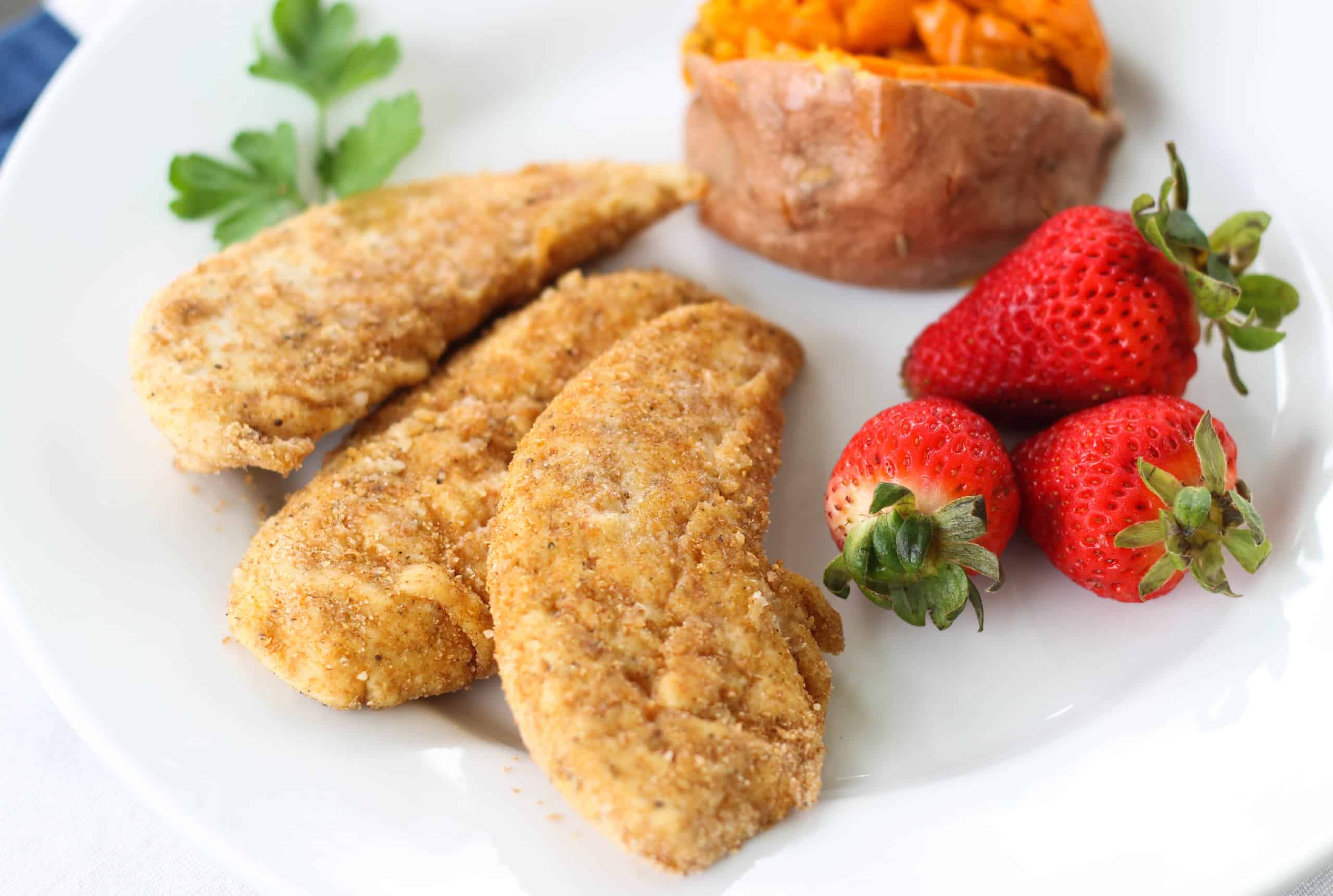 Three oven baked chicken tenders on a plate with strawberries and a baked sweet potato.