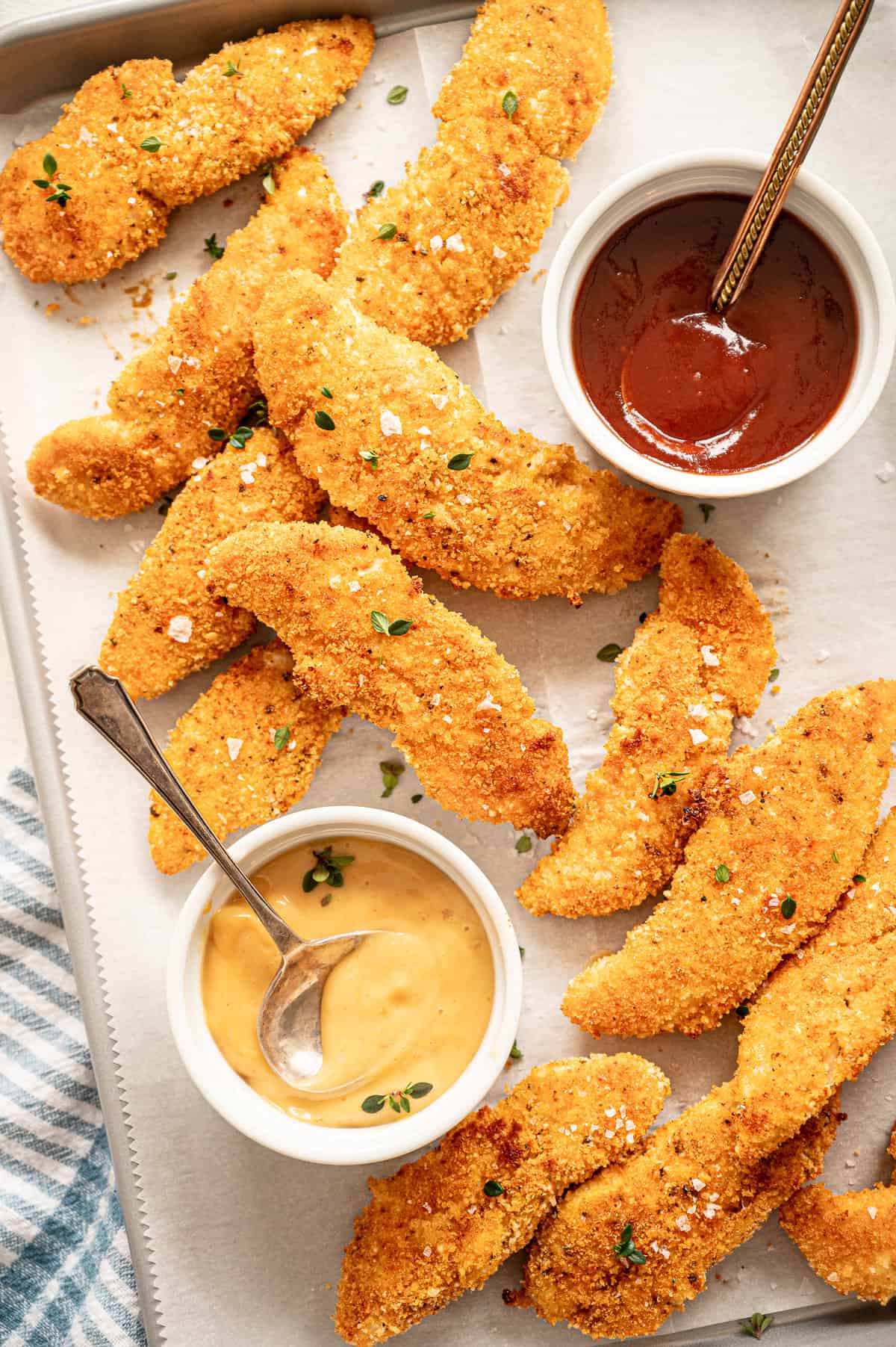 Parmesan chicken tenders on a parchment lined baking sheet with small bowls of honey mustard and ketchup on the side.